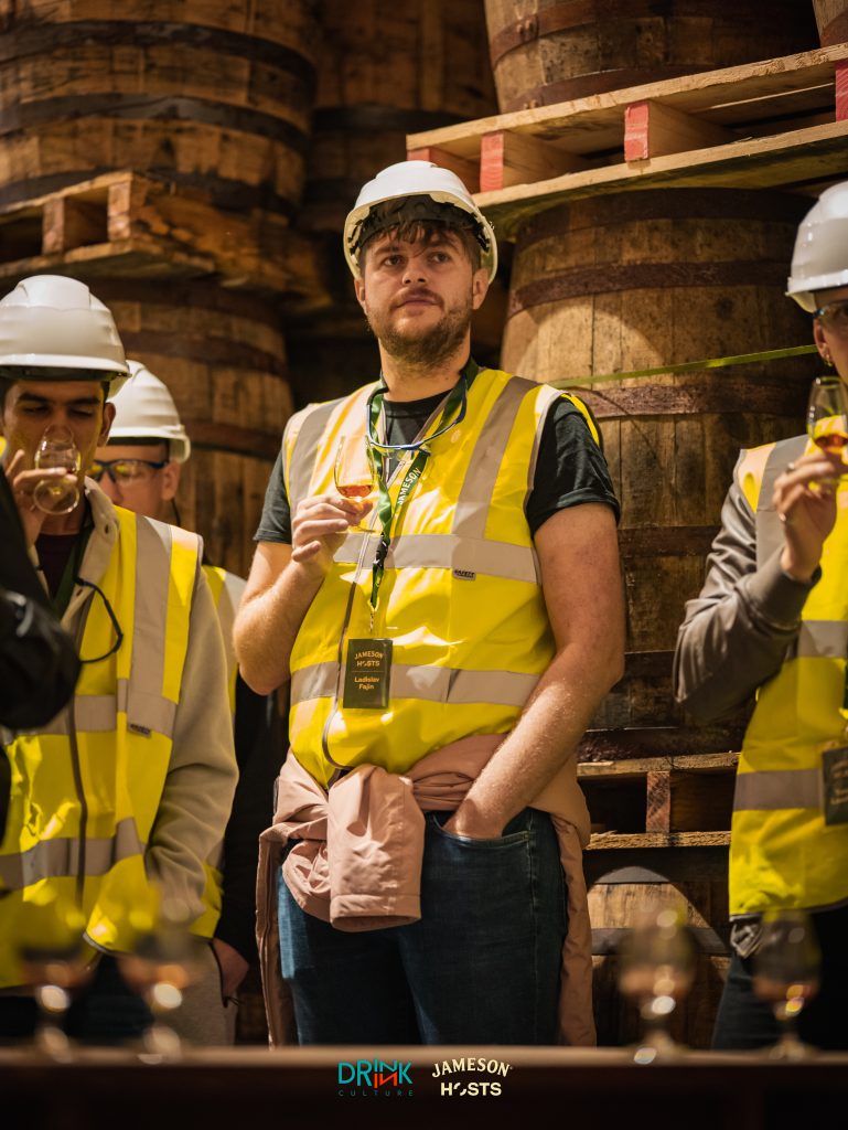 Photo of man standing with a small glass of whiskey surrounded by whiskey barrels in the Jameson distillery in Midleton.